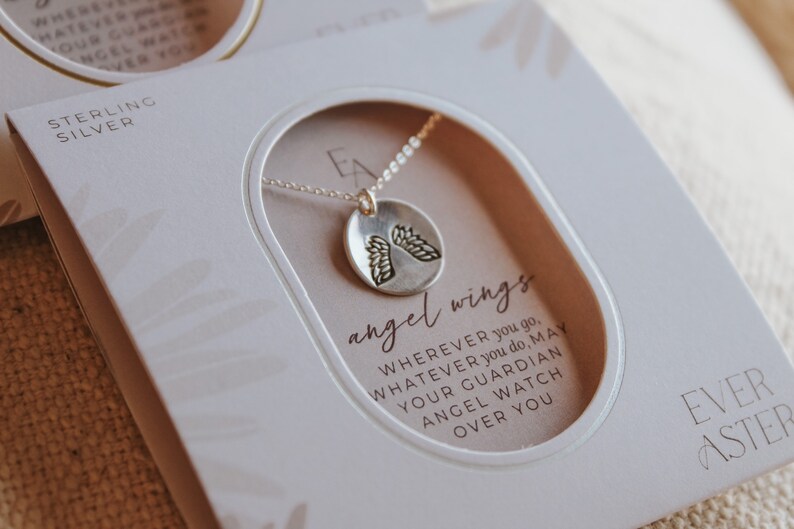 Puede incluir: Una caja blanca con un collar de plata con un peque&ntilde;o colgante redondo con alas de &aacute;ngel. La caja tiene el texto "sterling silver" y "angel wings". El texto en la caja dice: "Dondequiera que vayas, lo que sea que hagas, que tu &aacute;ngel de la guarda te cuide." La caja tambi&eacute;n tiene el texto "Ever After".