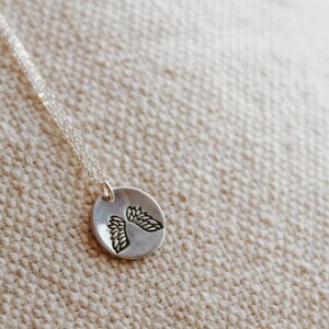 Puede incluir: Un collar de plata con un colgante redondo que presenta un dise&ntilde;o detallado de dos alas de &aacute;ngel.