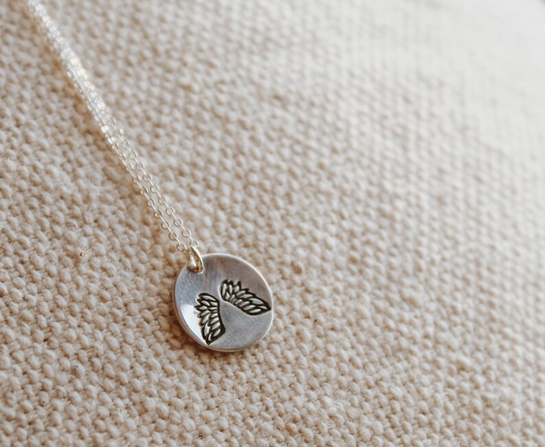 Puede incluir: Un collar de plata con un colgante redondo que presenta un dise&ntilde;o detallado de dos alas de &aacute;ngel.