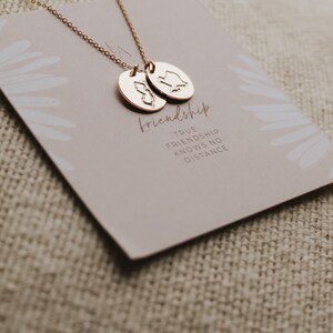 May include: A gold-coloured necklace with two oval pendants, each featuring a state outline. The necklace rests on a card with the word "friendship" and the phrase "TRUE FRIENDSHIP KNOWS NO DISTANCE". The background is a neutral beige.