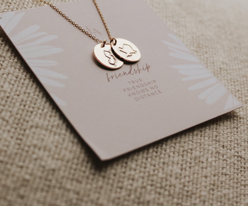May include: A gold-coloured necklace with two oval pendants, each featuring a state outline. The necklace rests on a card with the word "friendship" and the phrase "TRUE FRIENDSHIP KNOWS NO DISTANCE". The background is a neutral beige.
