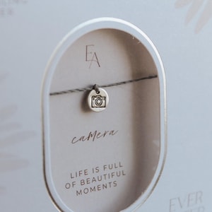 Peut inclure: Une carte blanche avec un cordon gris et un charme de caméra argenté. La carte dit "camera" et "La vie est pleine de beaux moments".