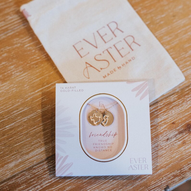 May include: A gold-filled necklace with two pendant charms, one shaped like Texas and the other like New Jersey, presented in a box with the words "friendship" and "true friendship knows no distance." A beige bag with the words "Ever Aster" is in the background.