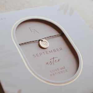 May include: A gray string bracelet with a gold charm featuring a flower design. The card reads "SEPTEMBER aster LOVE and PATIENCE".
