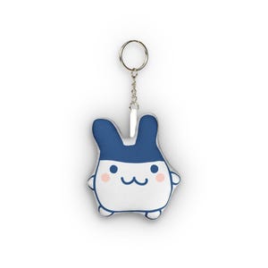 Puede incluir: Un llavero de peluche con forma de criatura blanca estilizada, parte superior azul oscuro, dos orejas altas, ojos negros pequeños, boca ondulada y mejillas rosadas circulares, unido a una anilla de metal plateado mediante una cadena.