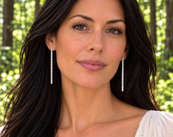 May include: A pair of long, slender, silver-toned bar earrings featuring a textured, hammered surface. The earrings hang vertically from the earlobes, showcasing a minimalist, straight design with a reflective metallic finish.