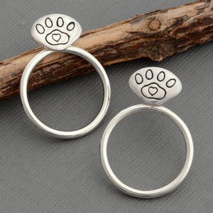 May include: A pair of silver-colored hoop earrings. Each earring features a paw print design with a heart in the center. The earrings are circular and have a polished finish. The background is a neutral gray.