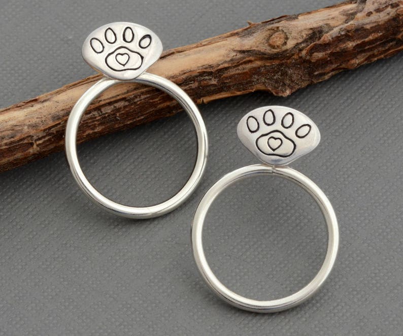 May include: A pair of silver-colored hoop earrings. Each earring features a paw print design with a heart in the center. The earrings are circular and have a polished finish. The background is a neutral gray.