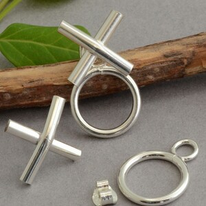 May include: A collection of silver-colored jewelry components, including two cross-shaped pieces, two circular rings, and a small earring back. The pieces are displayed on a neutral gray surface with a wooden branch and green leaf.