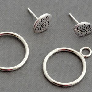 May include: A set of silver-colored jewelry components, including two circular rings with small loops and two paw print stud earrings. The paw prints have a heart in the center. The items are displayed on a gray surface.