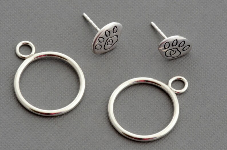 May include: A set of silver-colored jewelry components, including two circular rings with small loops and two paw print stud earrings. The paw prints have a heart in the center. The items are displayed on a gray surface.