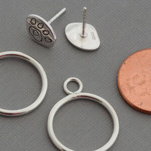 May include: Silver-toned jewelry components, including two circular pendant frames with small loops, and a pair of paw print stud earrings. A copper penny is included for size reference. The items are displayed on a gray surface.