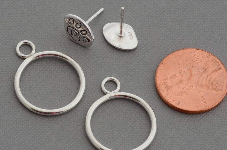 May include: Silver-toned jewelry components, including two circular pendant frames with small loops, and a pair of paw print stud earrings. A copper penny is included for size reference. The items are displayed on a gray surface.