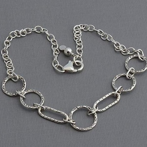 May include: A silver-toned necklace featuring a mix of chain links and textured oval and round links. The necklace has a lobster clasp closure. The jewelry is displayed on a gray background.