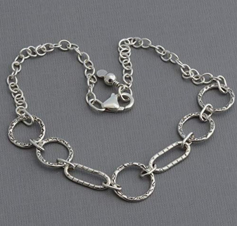 May include: A silver-toned necklace featuring a mix of chain links and textured oval and round links. The necklace has a lobster clasp closure. The jewelry is displayed on a gray background.