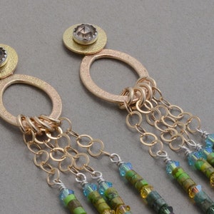 May include: Gold earrings with a round pendant and a chain of small gold rings. The pendant is adorned with a brown gemstone. The chain is attached to a series of green, blue, and purple beads.