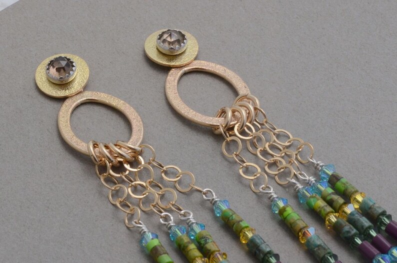 May include: Gold earrings with a round pendant and a chain of small gold rings. The pendant is adorned with a brown gemstone. The chain is attached to a series of green, blue, and purple beads.