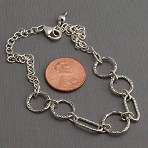 May include: A silver-colored bracelet with textured circular and oval links, and a lobster clasp. A penny is next to the bracelet for size comparison. The bracelet is on a gray surface.