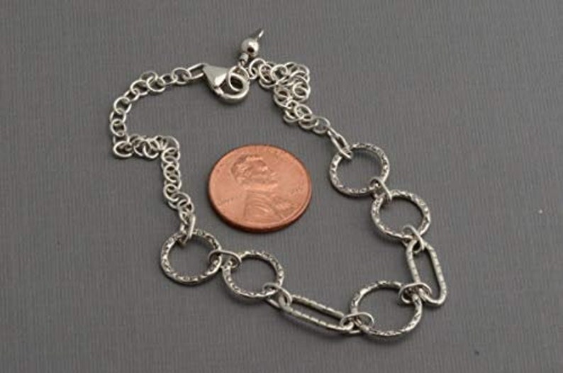 May include: A silver-colored bracelet with textured circular and oval links, and a lobster clasp. A penny is next to the bracelet for size comparison. The bracelet is on a gray surface.