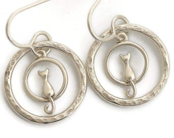 May include: A pair of silver-toned drop earrings featuring a small cat silhouette suspended within a larger, hammered-texture circular hoop. Each earring hangs from a simple curved wire hook, showcasing a polished metallic finish throughout.