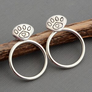 May include: A pair of silver-colored hoop earrings. Each earring features a paw print design with a heart in the center, set atop a circular hoop. The earrings are displayed on a wooden stick against a gray background.