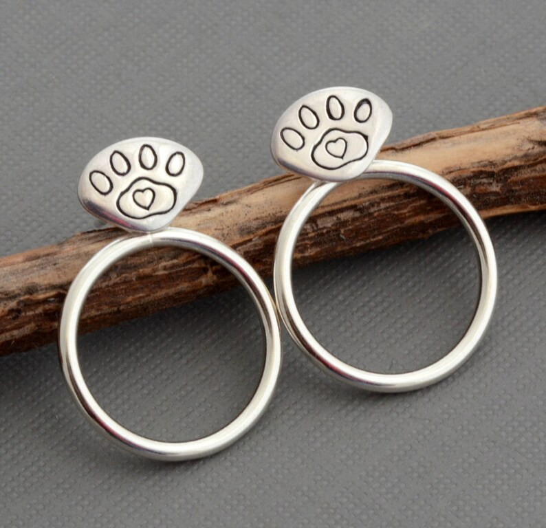 May include: A pair of silver-colored hoop earrings. Each earring features a paw print design with a heart in the center, set atop a circular hoop. The earrings are displayed on a wooden stick against a gray background.