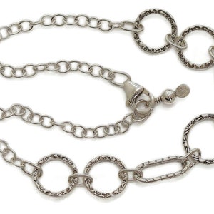 May include: Silver-toned necklace featuring a chain design with textured circular and oval links. The necklace has a lobster clasp closure and an adjustable chain. The decorative links have a detailed pattern. The jewelry is displayed against a white background.