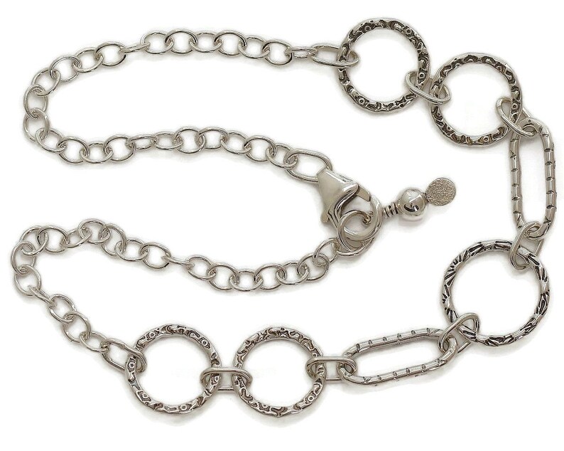 May include: Silver-toned necklace featuring a chain design with textured circular and oval links. The necklace has a lobster clasp closure and an adjustable chain. The decorative links have a detailed pattern. The jewelry is displayed against a white background.