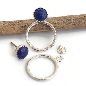 May include: A pair of silver earrings with a hammered texture, featuring a dark blue, round gemstone. One earring is a stud, the other is a hoop with a gemstone. A silver earring back is also present.