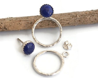 May include: A pair of silver earrings with a hammered texture, featuring a dark blue, round gemstone. One earring is a stud, the other is a hoop with a gemstone. A silver earring back is also present.