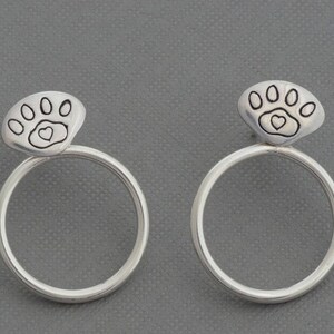 May include: A pair of silver hoop earrings. Each earring features a paw print design with a heart in the center. The earrings are set against a gray background.