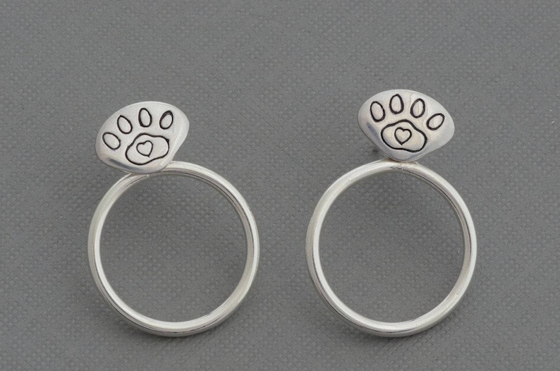 May include: A pair of silver hoop earrings. Each earring features a paw print design with a heart in the center. The earrings are set against a gray background.