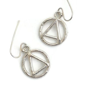May include: A pair of silver-toned earrings. Each earring features a circular design with a hammered texture, enclosing a triangular shape. The earrings have a simple hook closure and are displayed against a white background.