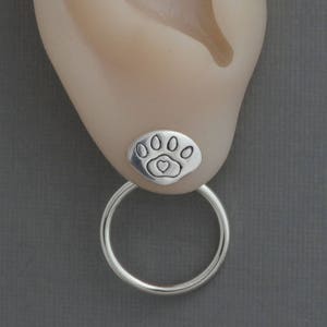 May include: A silver earring featuring a paw print design with a heart in the center. The earring has a small, round stud and a larger hoop that hangs below. The earring is shown on an ear.