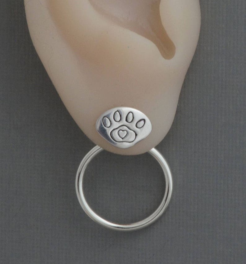 May include: A silver earring featuring a paw print design with a heart in the center. The earring has a small, round stud and a larger hoop that hangs below. The earring is shown on an ear.