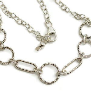 May include: A silver-colored necklace featuring a series of textured, open circles and oval links. The necklace has a chain with a lobster clasp closure. The design is simple and elegant, suitable for everyday wear.