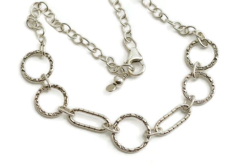 May include: A silver-colored necklace featuring a series of textured, open circles and oval links. The necklace has a chain with a lobster clasp closure. The design is simple and elegant, suitable for everyday wear.