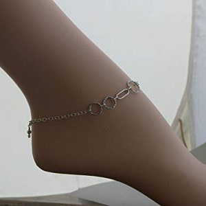 May include: A silver-colored anklet with a delicate chain and a series of connected circles and ovals. A small charm hangs near the ankle. The jewelry is worn on a light-skinned foot.