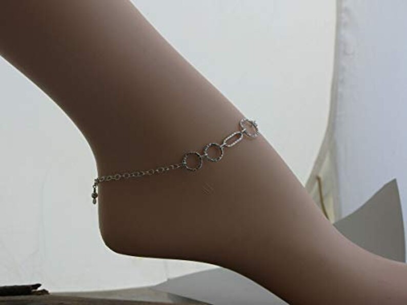 May include: A silver-colored anklet with a delicate chain and a series of connected circles and ovals. A small charm hangs near the ankle. The jewelry is worn on a light-skinned foot.