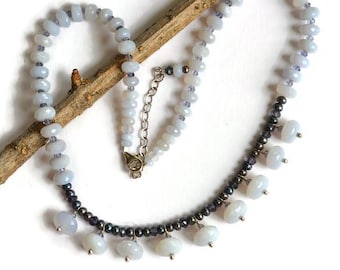 May include: A necklace featuring pale blue, disc-shaped beads interspersed with small, dark grey beads. The lower section includes dangling pale blue beads attached to a dark grey bead strand. A silver-toned metal chain and clasp are at the top.