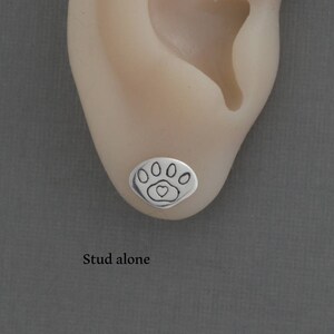 May include: A silver stud earring with a paw print design, featuring five oval shapes and a heart in the center. The earring is shown on an ear against a gray background. The text "Stud alone" is below the earring.