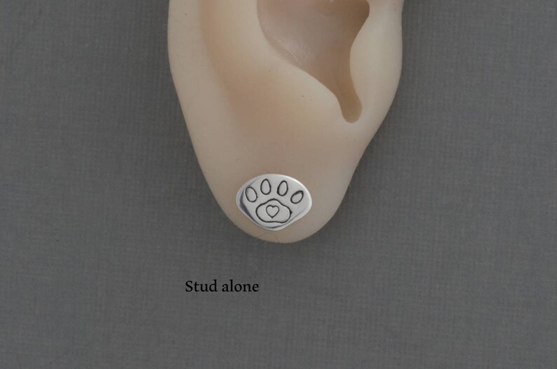 May include: A silver stud earring with a paw print design, featuring five oval shapes and a heart in the center. The earring is shown on an ear against a gray background. The text "Stud alone" is below the earring.
