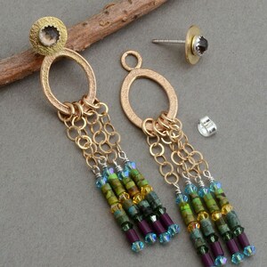 May include: Gold dangle earrings with a textured oval ring and a small gemstone at the top. The earrings feature multiple strands of gold chain and colorful beads in shades of green, blue, and purple.