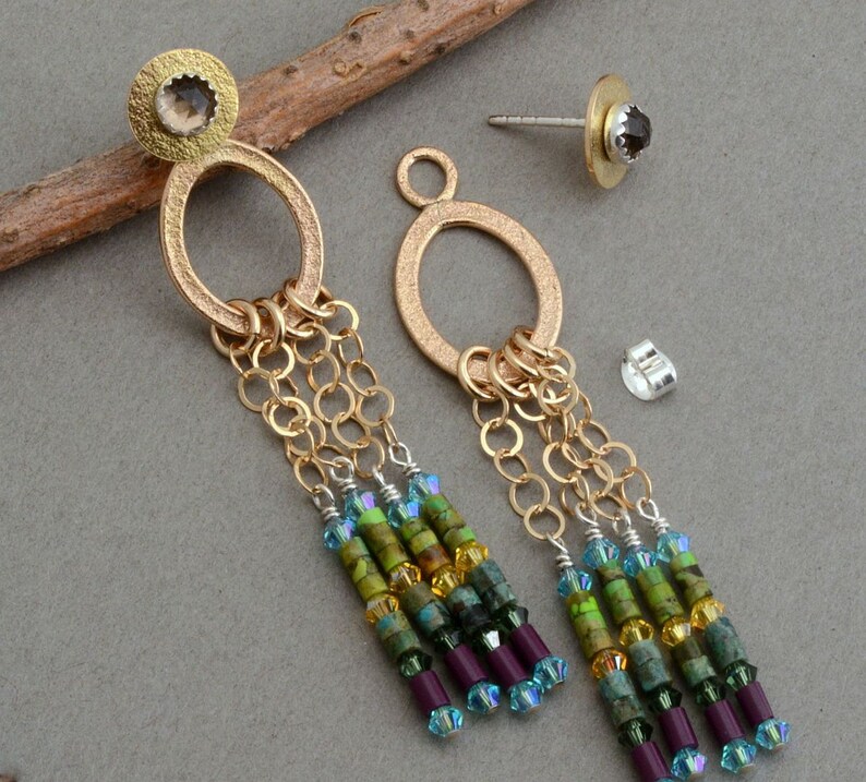May include: Gold dangle earrings with a textured oval ring and a small gemstone at the top. The earrings feature multiple strands of gold chain and colorful beads in shades of green, blue, and purple.