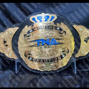 Könnte beinhalten: Ein gold-schwarzer Wrestling-Meisterschaftsgürtel. Der Gürtel trägt den Text "WOMENS TNA WRESTLING CHAMPION" in goldfarbenen Buchstaben. Der Gürtel hat ein blaues Kronen-Design oben und goldfarbene Akzente.