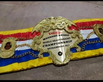 Może przedstawiać: Złoty pas mistrzowski w boksie z napisem "RING MAGAZINE AWARD WORLD HEAVYWEIGHT CHAMPIONSHIP MUHAMMAD ALI". Pas ma wielokolorową podstawę z tkaniny z czerwonymi, białymi, niebieskimi i żółtymi paskami.