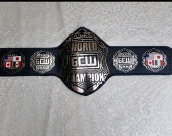 May include: A black wrestling championship belt with silver accents. The belt features the text "WORLD GCW CHAMPION" and includes emblems with flags of the United States, Canada, and Japan. The belt is displayed on a textured surface.