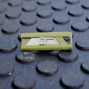 May include: An olive green utility knife with a silver blade and a gray button. The knife is rectangular and has a geometric design. The background is a dark gray surface with a repeating pattern of raised circles.