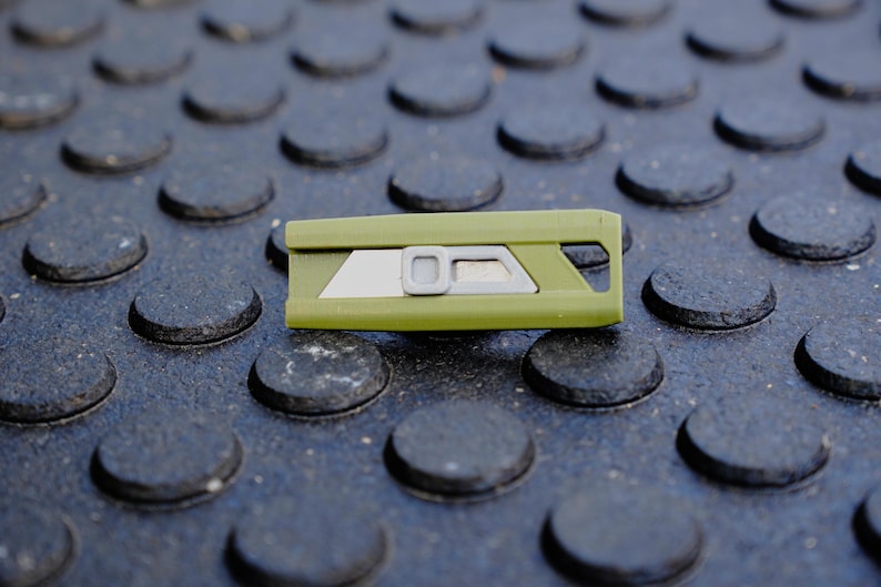 May include: An olive green utility knife with a silver blade and a gray button. The knife is rectangular and has a geometric design. The background is a dark gray surface with a repeating pattern of raised circles.