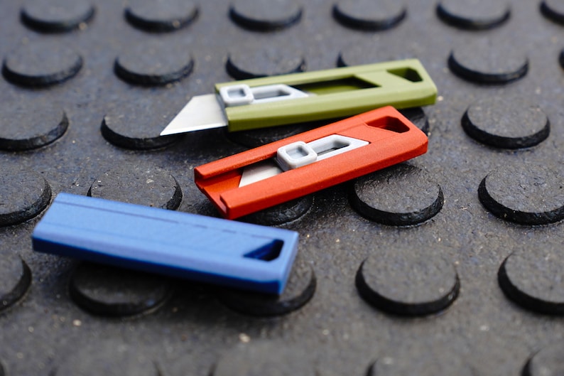 May include: Three utility knives in green, red, and blue. The green knife has its blade extended, while the red knife has its blade retracted. The blue knife is closed. All three knives have a gray slider and a silver blade.